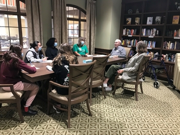 Students gather around a table to listen and learn from a retirement home resident.