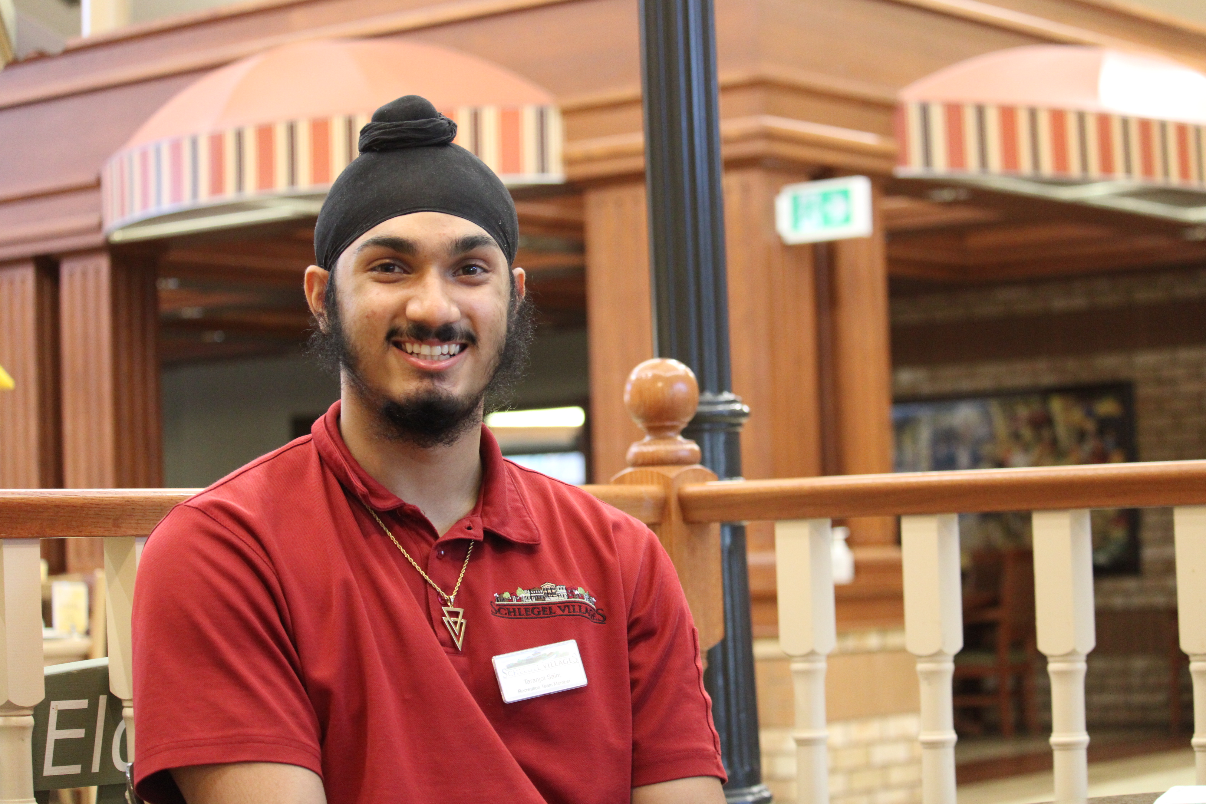 Taranjot stands alongside the railing in the Village along Main Street, smiling wide in his red Schlegel Villages shirt. 