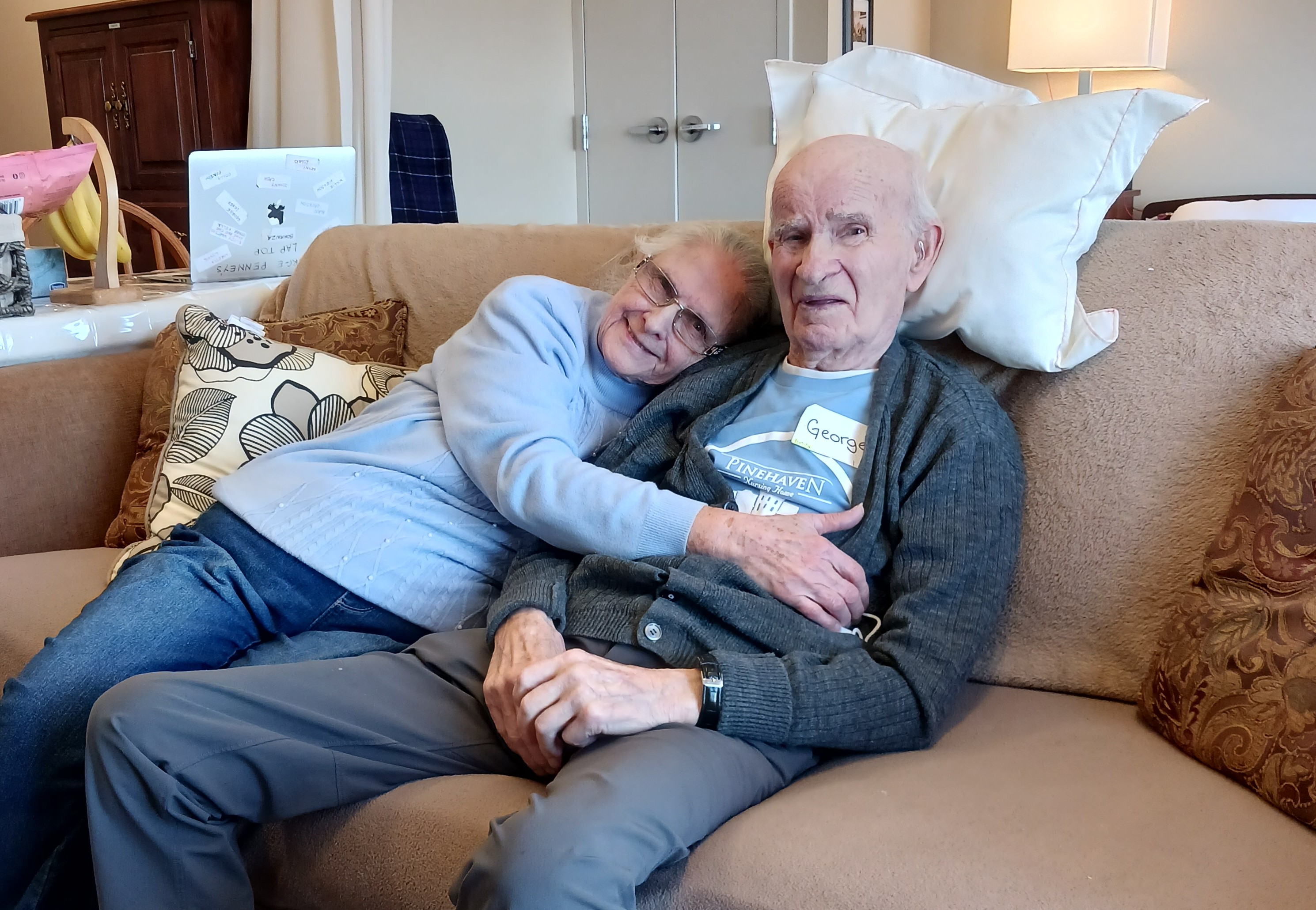 Ursula embraces George as they sit together on the couch in their comfortable suite. 