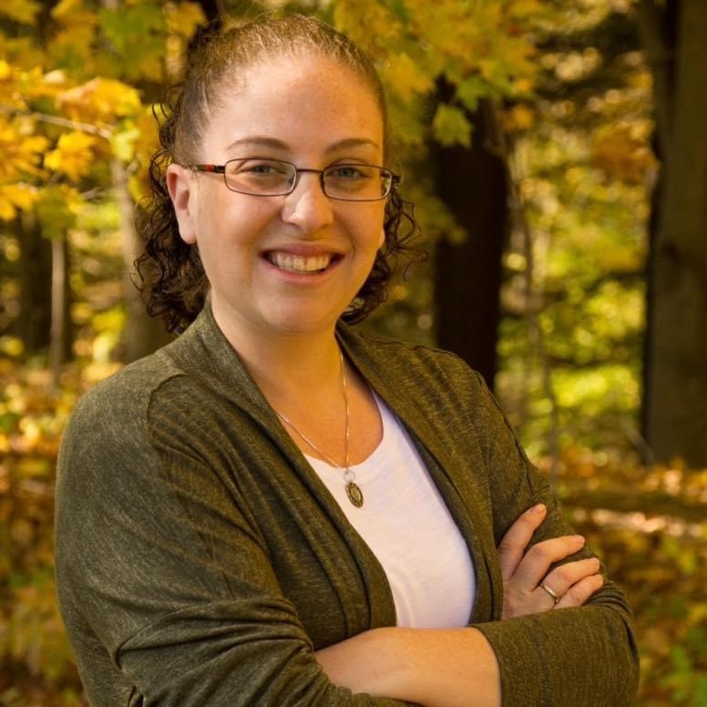 A photo of Emily Baker smiling under Autumn leaves. 