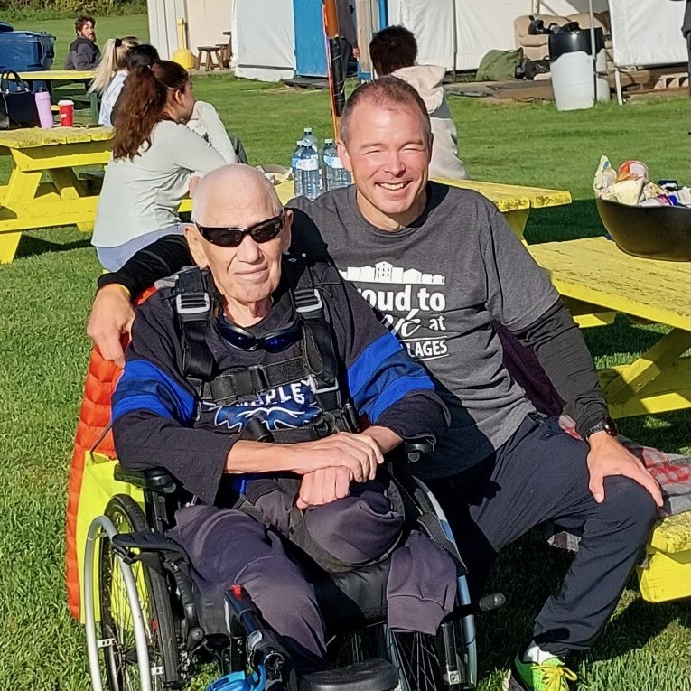 Bryce kneels alongside a resident in a wheelchair who is about to go skydiving. 