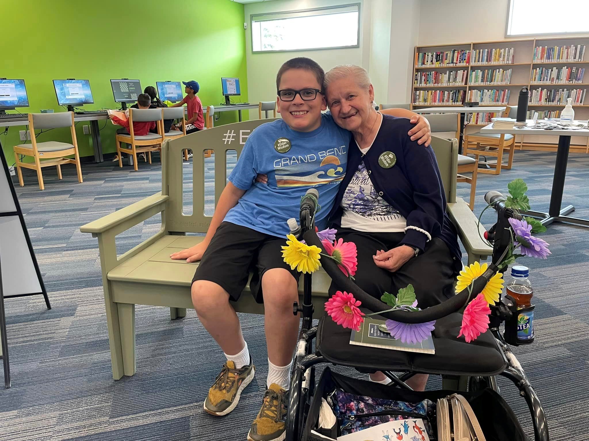 A young person and an older person share a smiling embrace upon the #ElderWisdom Green Bench.